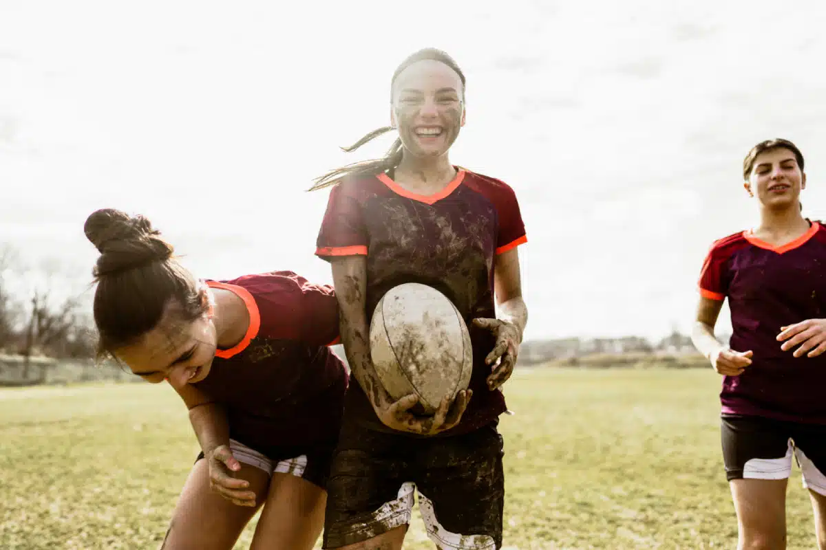 Three young women wearing rugby uniforms, muddy and smiling, stand together on a grassy field. One holds a rugby ball while the others laugh, enjoying a playful and energetic moment outdoors.