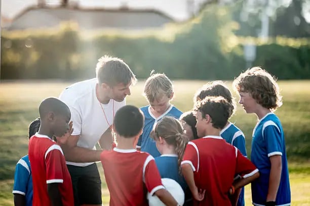 A coach kneels and talks to a group of young soccer players wearing red and blue jerseys. The kids are gathered closely in a huddle on a grassy field, listening attentively during a sunny day.