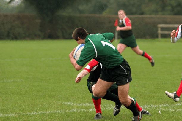 A rugby player in a green jersey runs with the ball while an opponent in a red jersey attempts to tackle him on a grassy field. Other players are visible in the background.