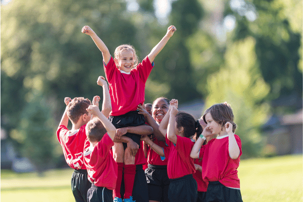 A group of kids in red shirts celebrate outdoors, lifting a smiling girl with raised arms above their heads. The children are joyful, and the scene is set in a sunny, green park.