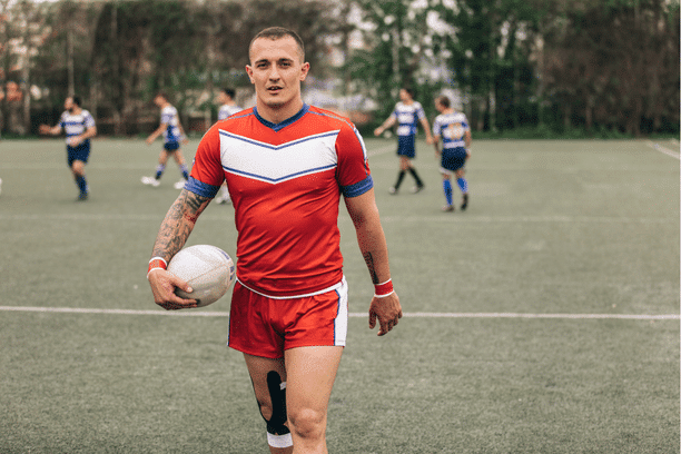 A rugby player in a red and white uniform holds a rugby ball and walks on a grassy field, with teammates and opponents blurred in the background.