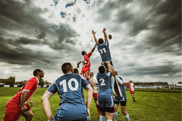Rugby players in blue and red uniforms jump and reach for the ball during a line-out on a grassy field under a dramatic, cloudy sky. Spectators and buildings are visible in the background.