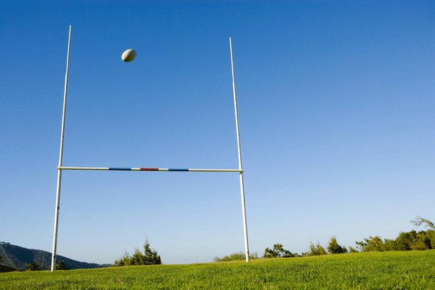 A rugby ball is flying through the air towards a goalpost on a grassy field under a clear blue sky. Trees and distant hills are visible in the background.