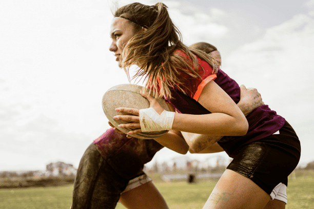 A female rugby player runs with the ball while being tackled by an opponent during a game on a grassy field. Both athletes appear focused and determined.