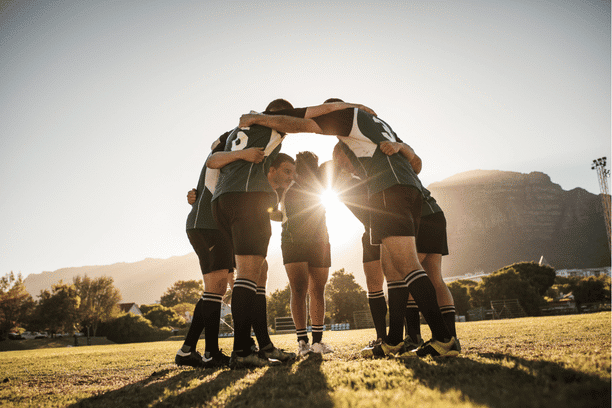 A group of rugby players in uniforms huddle together on a grassy field at sunset, with sunlight shining through their circle and mountains visible in the background.