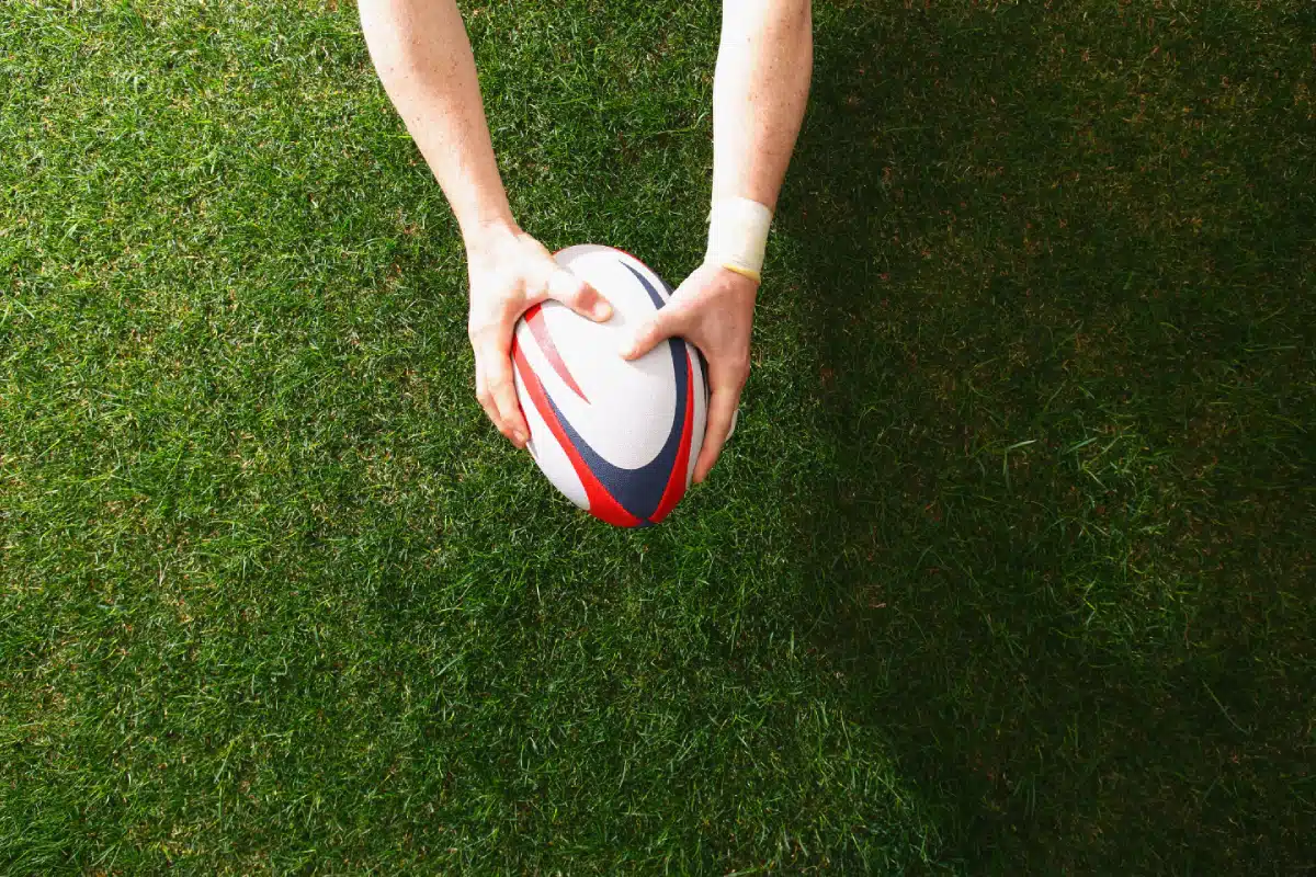 A person’s hands are placing a white rugby ball with red and blue stripes on green grass, viewed from above. The field is well-manicured and the person's arms are visible from the elbows down.
